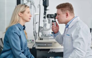 Eine Frau sitzt in einer Augenarztpraxis und wird von einem Arzt untersucht, der mit einem Ophthalmoskop in ihre Augen schaut. Im Hintergrund ist medizinisches Untersuchungsgerät zur Augenuntersuchung zu sehen.