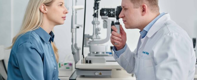 Eine Frau sitzt in einer Augenarztpraxis und wird von einem Arzt untersucht, der mit einem Ophthalmoskop in ihre Augen schaut. Im Hintergrund ist medizinisches Untersuchungsgerät zur Augenuntersuchung zu sehen.