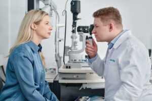 Eine Frau sitzt in einer Augenarztpraxis und wird von einem Arzt untersucht, der mit einem Ophthalmoskop in ihre Augen schaut. Im Hintergrund ist medizinisches Untersuchungsgerät zur Augenuntersuchung zu sehen.