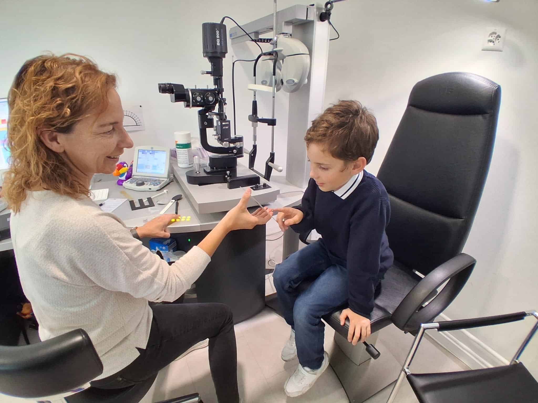 Ein Junge sitzt auf einem schwarzen Stuhl, eine Frau zeigt ihm eine graue Karte mit Mustern. Der Junge deutet mit dem Finger auf die Karte. Beide befinden sich in einem augenoptischen Raum mit technischen Geräten, einem Monitor und verschiedenen Hilfsmitteln auf dem Tisch.