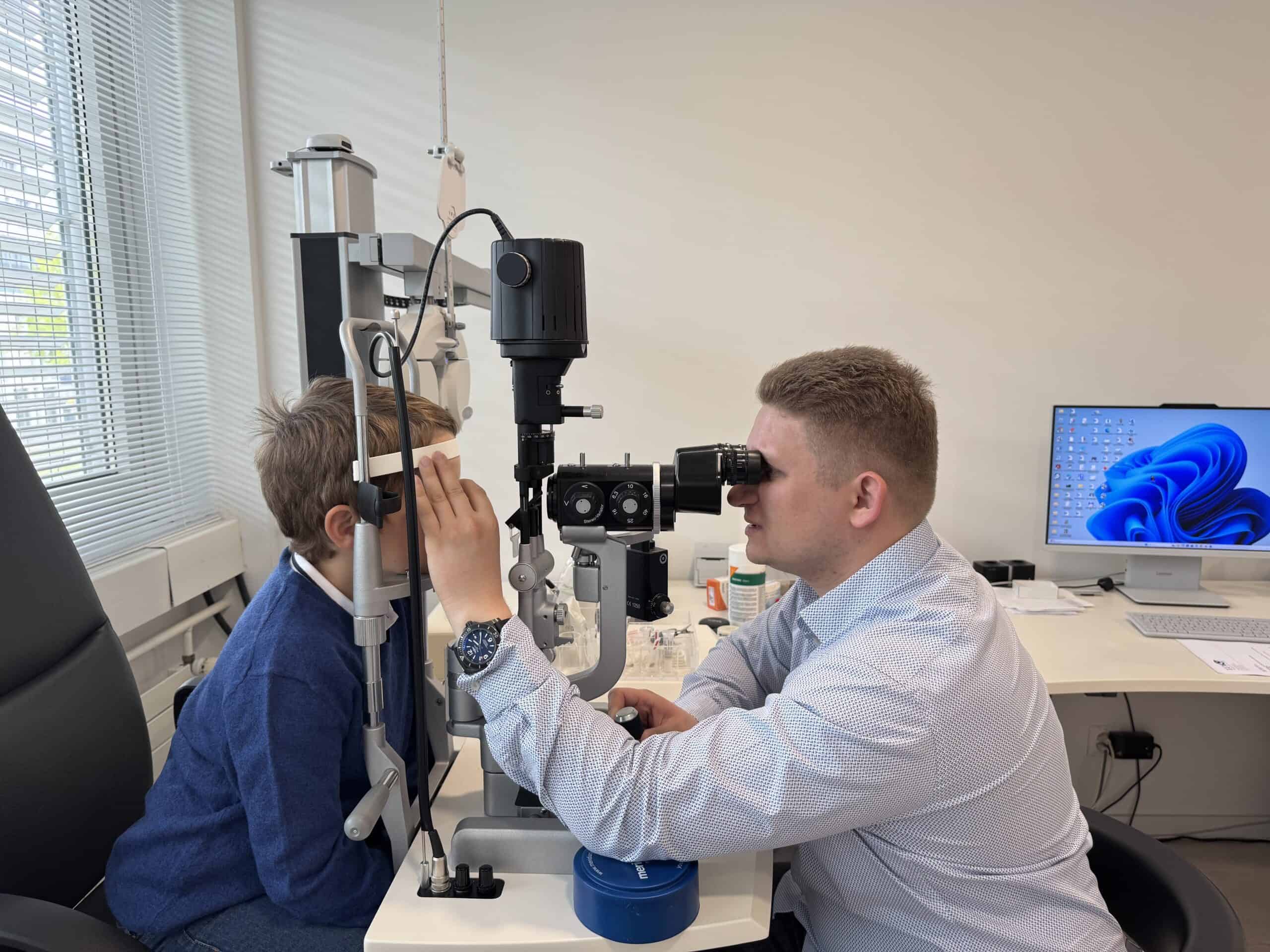 Ein Junge verdeckt ein Auge mit einem Papierstreifen, während der Mann ihn durch das Spaltlampenmikroskop untersucht.