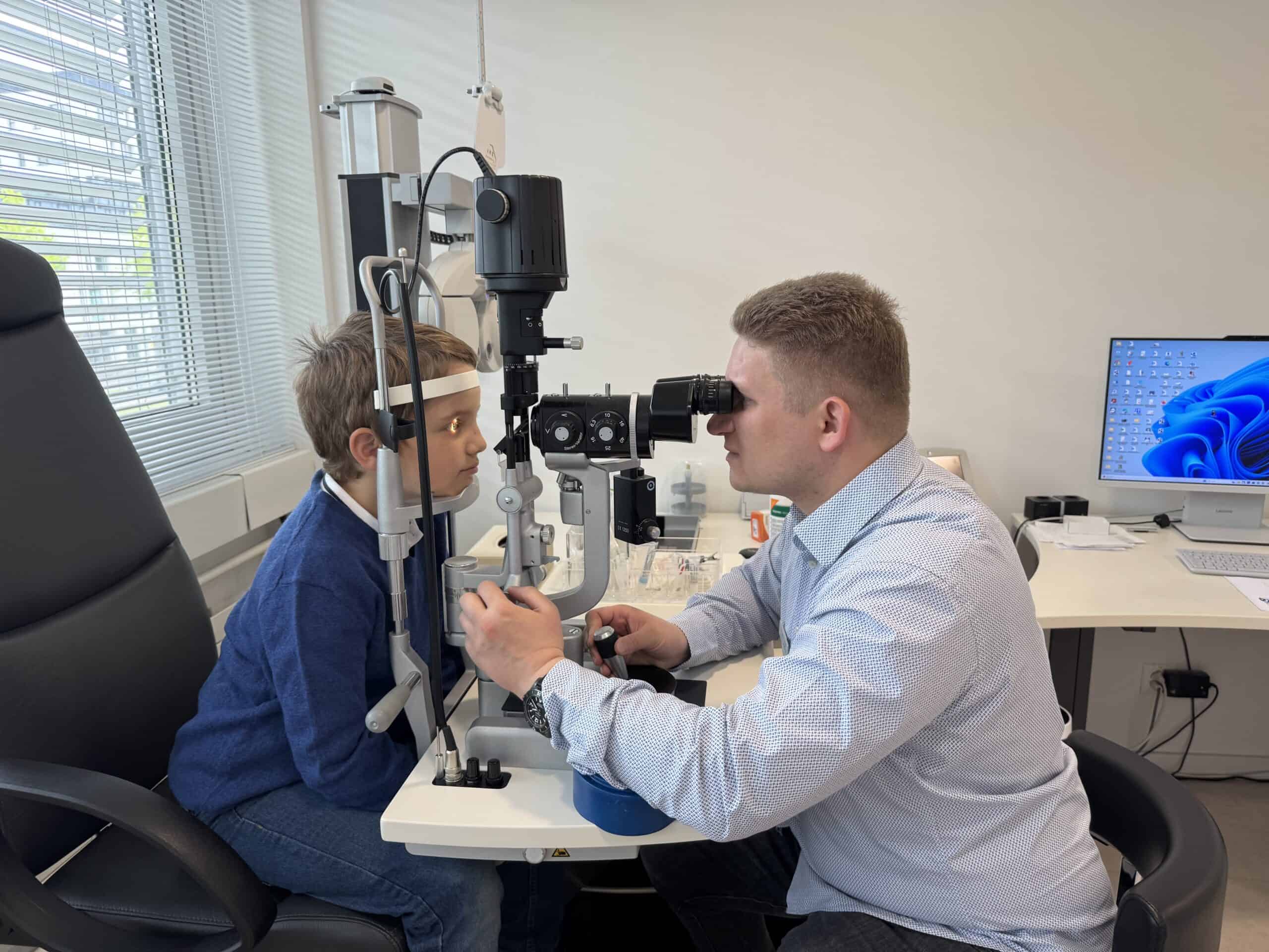 Ein Junge sitzt an einem Spaltlampenmikroskop, trägt ein Stirnband zur Fixierung des Kopfes und schaut geradeaus. Ein Mann in hellem Hemd untersucht ihn durch das Mikroskop. Im Hintergrund stehen ein Bildschirm mit Windows-Desktop, ein Schreibtisch und verschiedene medizinische Geräte.