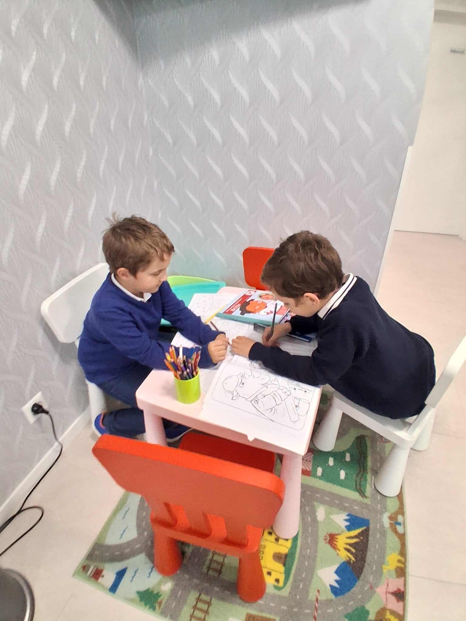 Zwei Jungen sitzen an einem Kindertisch mit bunten Stühlen und malen in Ausmalbüchern. Auf dem Tisch stehen ein Becher mit Buntstiften und verschiedene Malutensilien. Der Boden ist mit einem Spielteppich mit Straßenmotiv bedeckt.