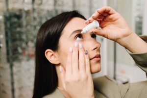 Nahaufnahme einer Person mit langen dunklen Haaren, die sich mit der rechten Hand Augentropfen ins rechte Auge verabreicht. Der Kopf ist leicht nach hinten geneigt und die Blickrichtung nach oben gerichtet. Die linke Hand zieht das untere Augenlid leicht nach unten. Die Fingernägel sind manikürt, einige mit hellem Lack, einer mit Zebramuster. Die Person trägt eine beigefarbene Kleidung. Im Hintergrund unscharf erkennbare Regale mit Brillenfassungen.