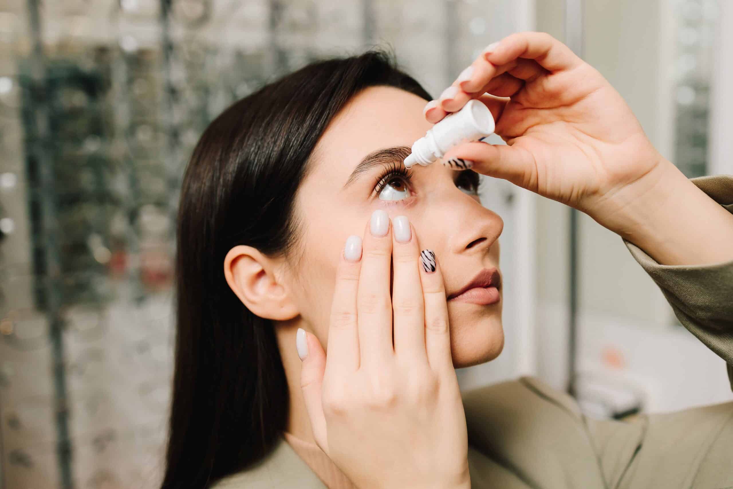 Nahaufnahme einer Person mit langen dunklen Haaren, die sich mit der rechten Hand Augentropfen ins rechte Auge verabreicht. Der Kopf ist leicht nach hinten geneigt und die Blickrichtung nach oben gerichtet. Die linke Hand zieht das untere Augenlid leicht nach unten. Die Fingernägel sind manikürt, einige mit hellem Lack, einer mit Zebramuster. Die Person trägt eine beigefarbene Kleidung. Im Hintergrund unscharf erkennbare Regale mit Brillenfassungen.