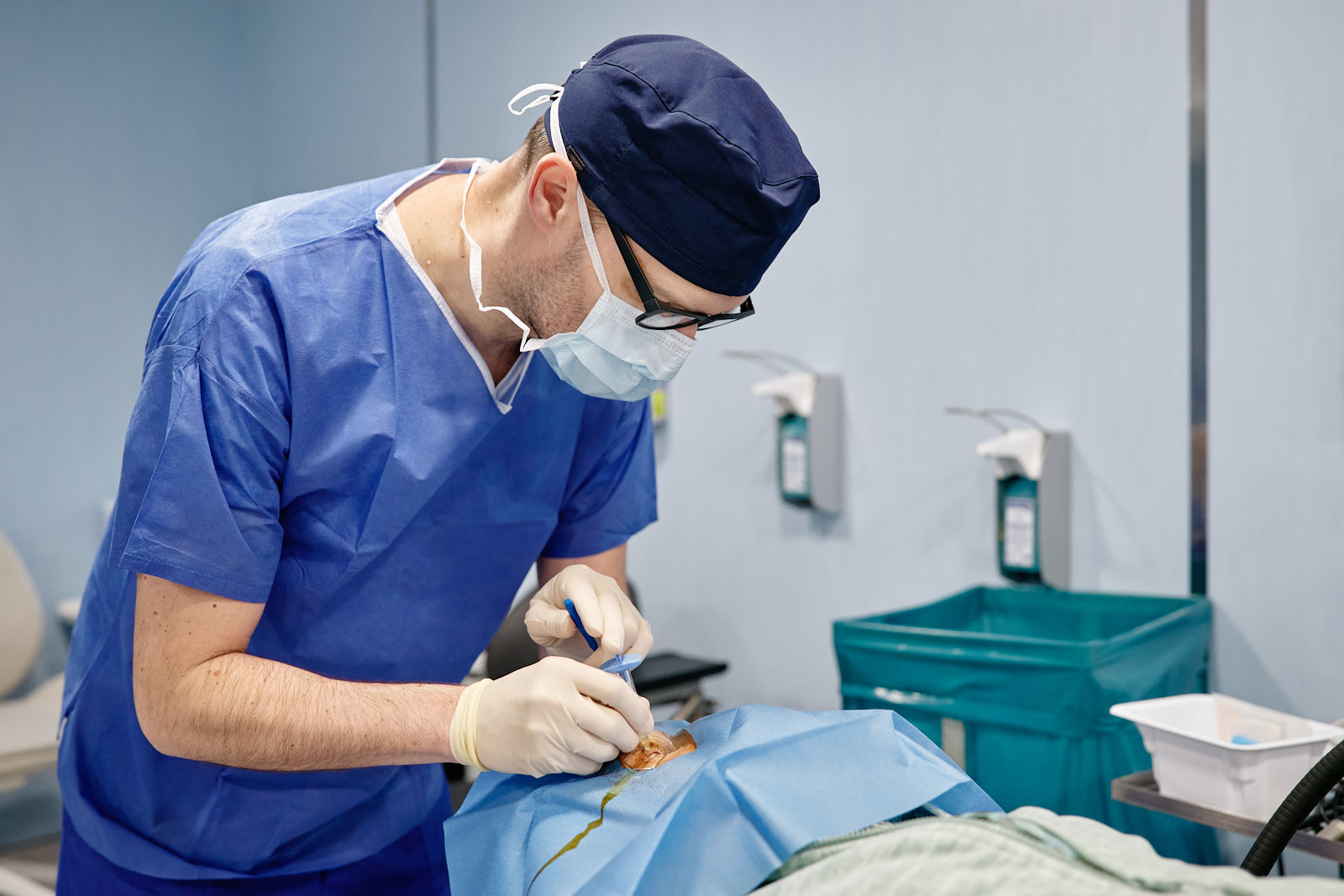 Person in blauer OP-Kleidung mit Haube, Brille, Mundschutz und Handschuhen beugt sich über einen abgedeckten Bereich und arbeitet mit einem medizinischen Instrument. Im Hintergrund sind helle Wände, Desinfektionsspender und ein türkisfarbener Behälter zu sehen.